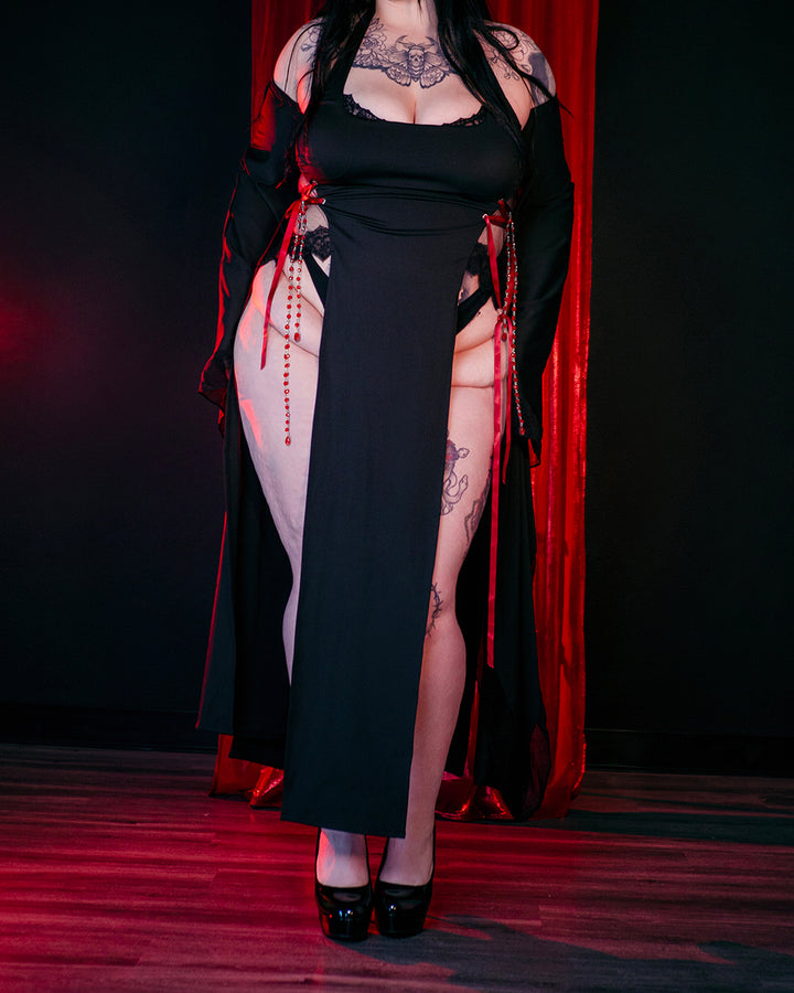 Witching Hour Dress by MOEFLAVOR, a dramatic black gown with oversized sleeves, open hip slits, red ribbon lace at the waist, and red crystal chain details for a magical allure